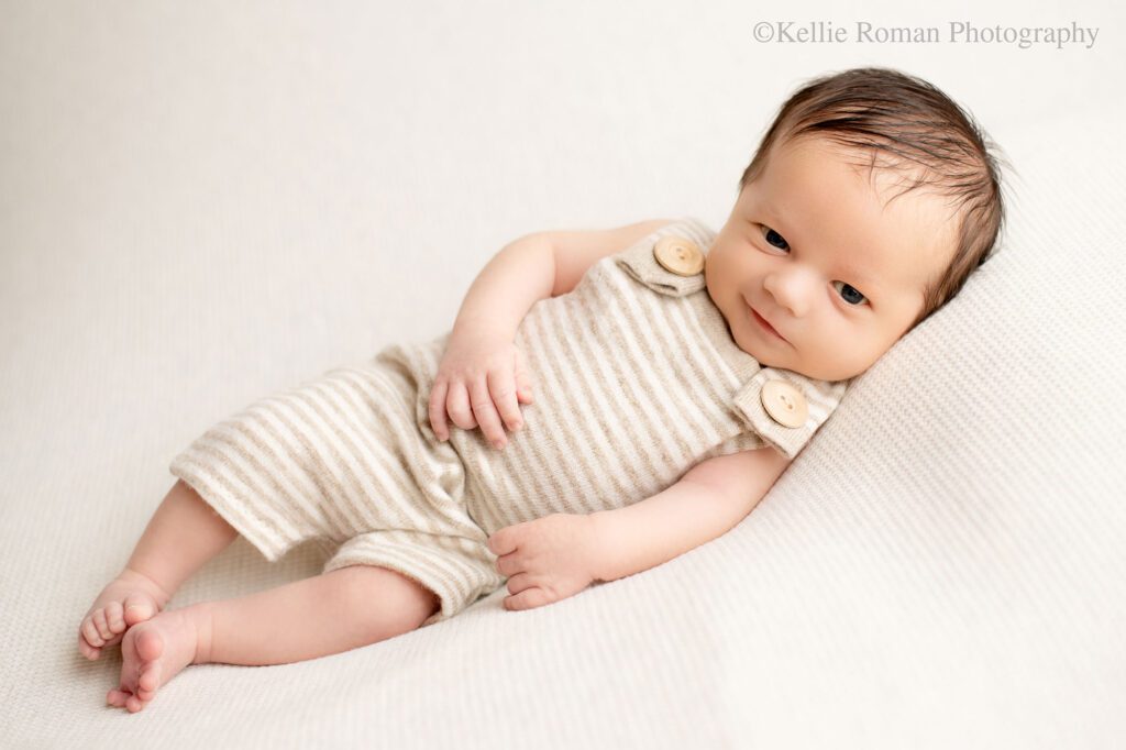 milwaukee newborn portraits. a newborn boy is laying on his side on cream fabric. he has a striped cream romper on with big wood buttons. he's awake and smirking at the camera. 