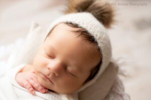 milwaukee newborn portraits. a newborn boy is sleeping with a cream hat on with tan pom pom. its a close up image of his face. he's in a cream swaddle with one hand sticking out.