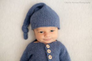 milwaukee newborn portraits. newborn boy laying on his back on top of cream fabric. he's awake and making a goofy smile. he's wearing a blue knit romper with wood buttons. he has a matching sleepy hat on.