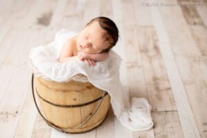 milwaukee newborn portraits. newborn boy sleeping in a wood bucket. his hands are under his chin, and the bucket is filled with cream layered fabrics. the baby boy has dark brown hair.