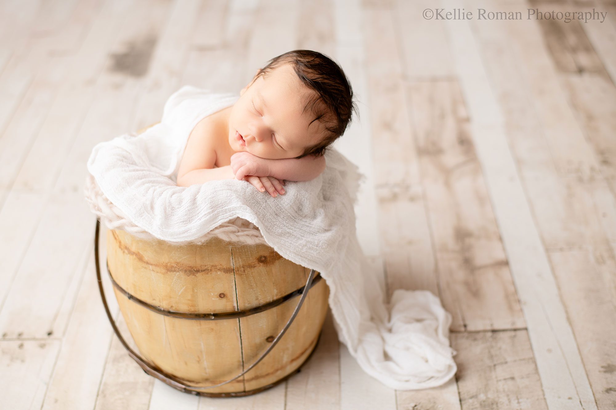 milwaukee newborn portraits. a newborn boy is sleeping in a wood bucket. his chin is resting on his hands. the bucket is stuffed with cream furs and layering fabric. 