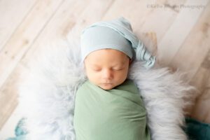 Milwaukee newborn portraits. newborn boy is sleeping in a wood basket filled with blue fur. he has a green swaddle on and a light blue sleepy hat.