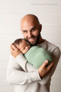 milwaukee newborn portraits. a father is in milwaukee photo studio holding his newborn son. the backdrop is white wood, the father has a cream shirt on. the newborn is in a green swaddle. the father is looking and smiling.
