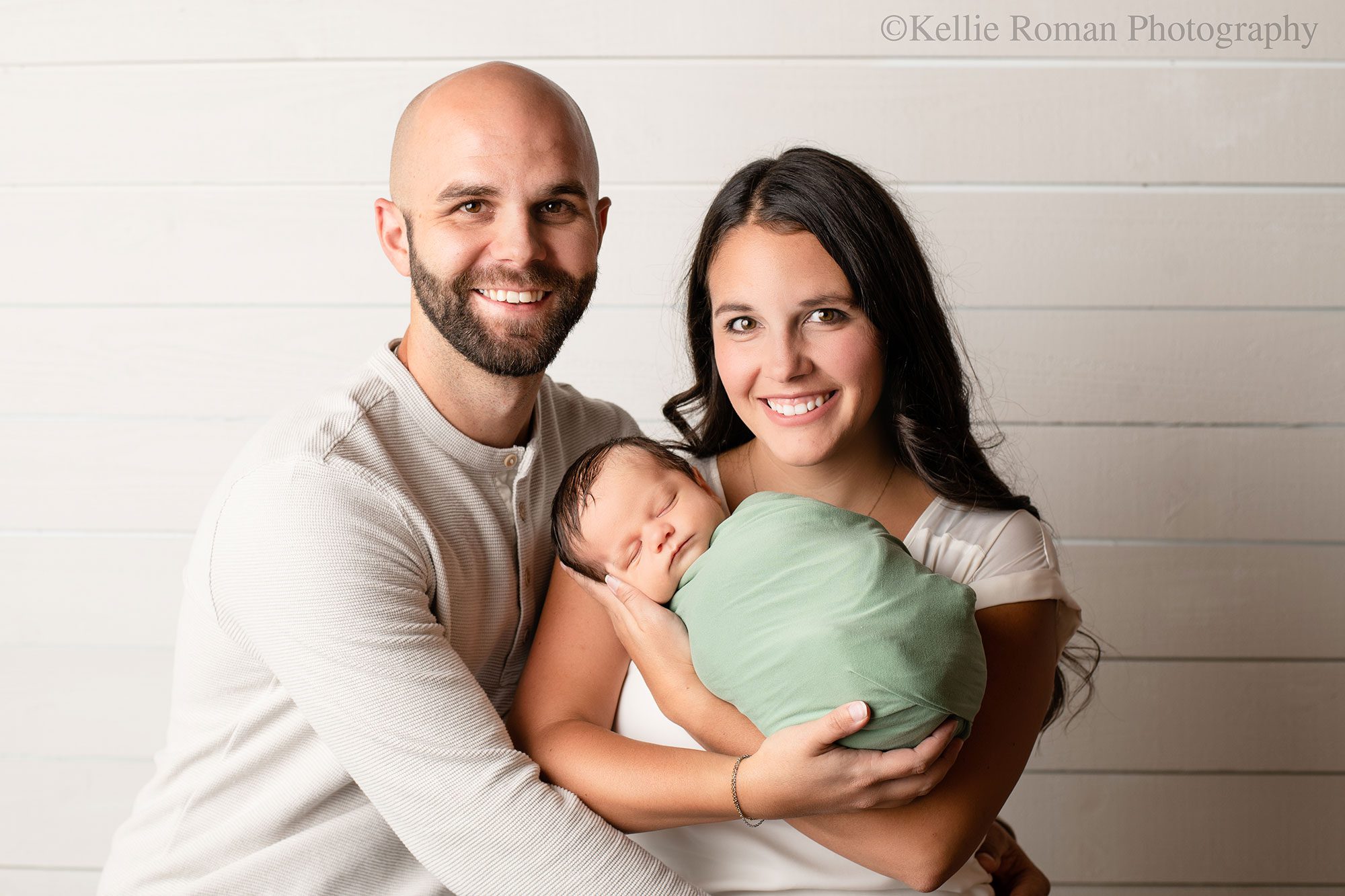 milwaukee newborn portraits. a family of three is in milwaukee photo studio smiling at camera. mom is holding her newborn boy who is wrapped in a green swaddle. 