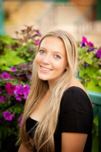 milwaukee senior photographer. high school senior girl headshot. she has a black top on with long blonde hair and blue eyes. there are green and purple flowers behind her.
