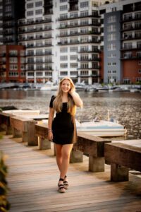 milwaukee senior photographer. high school senior girl standing on wood walkway along milwaukee river. girl has long blonde hair and is running her hand through it. she has a short black dress on and black sandals. there are tall condo buildings behind her.