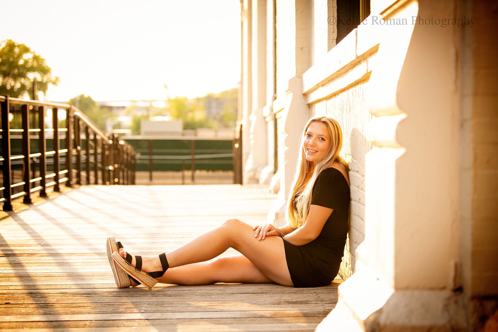 milwaukee senior photographer. high school senior girl sitting on wood walkway resting her back against cream brick building. girl has long blonde hair and is wearing a short black dress with black sandals.