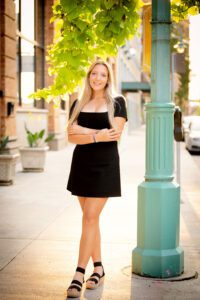 milwaukee senior photographer. high school senior girl standing on sidewalk next to teal light pole in milwaukee downtown. she has a short black dress on with black sandals. senior has long blonde hair, and is crossing her arms. sun is setting behind her, and a green vine is hanging from the lightpole.