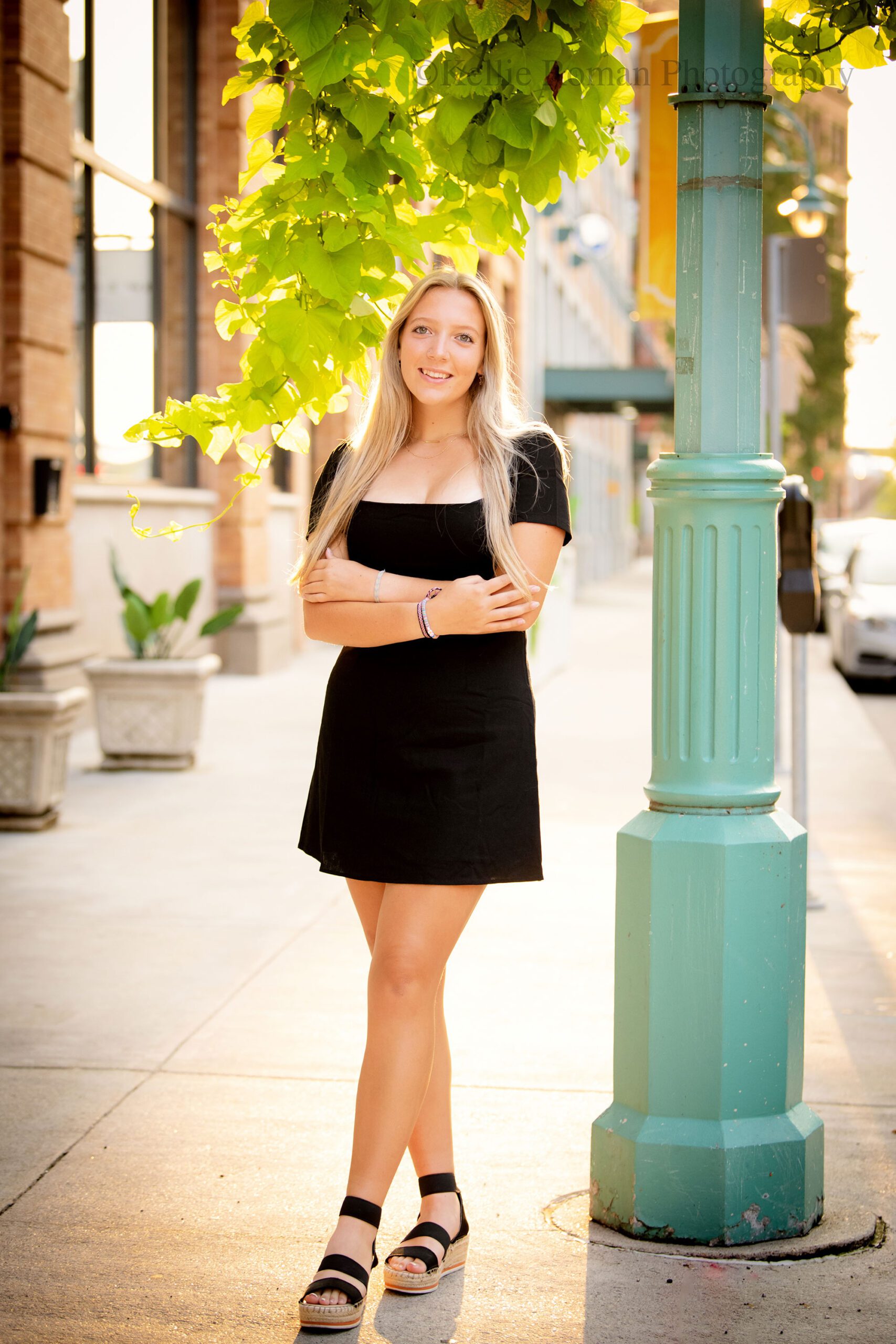 milwaukee senior photographer. high school senior girl is standing in Downtown Milwaukee next to teal light pole that has green vines hanging. she has black sandals on and a black short dress. she has long blonde hair. 