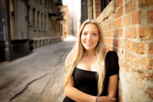 Milwaukee senior photographer. high school senior girl wearing short black dress has long blonde hair and blue eyes. she's leaning against cream colored old brick in milwaukee downtown ally.