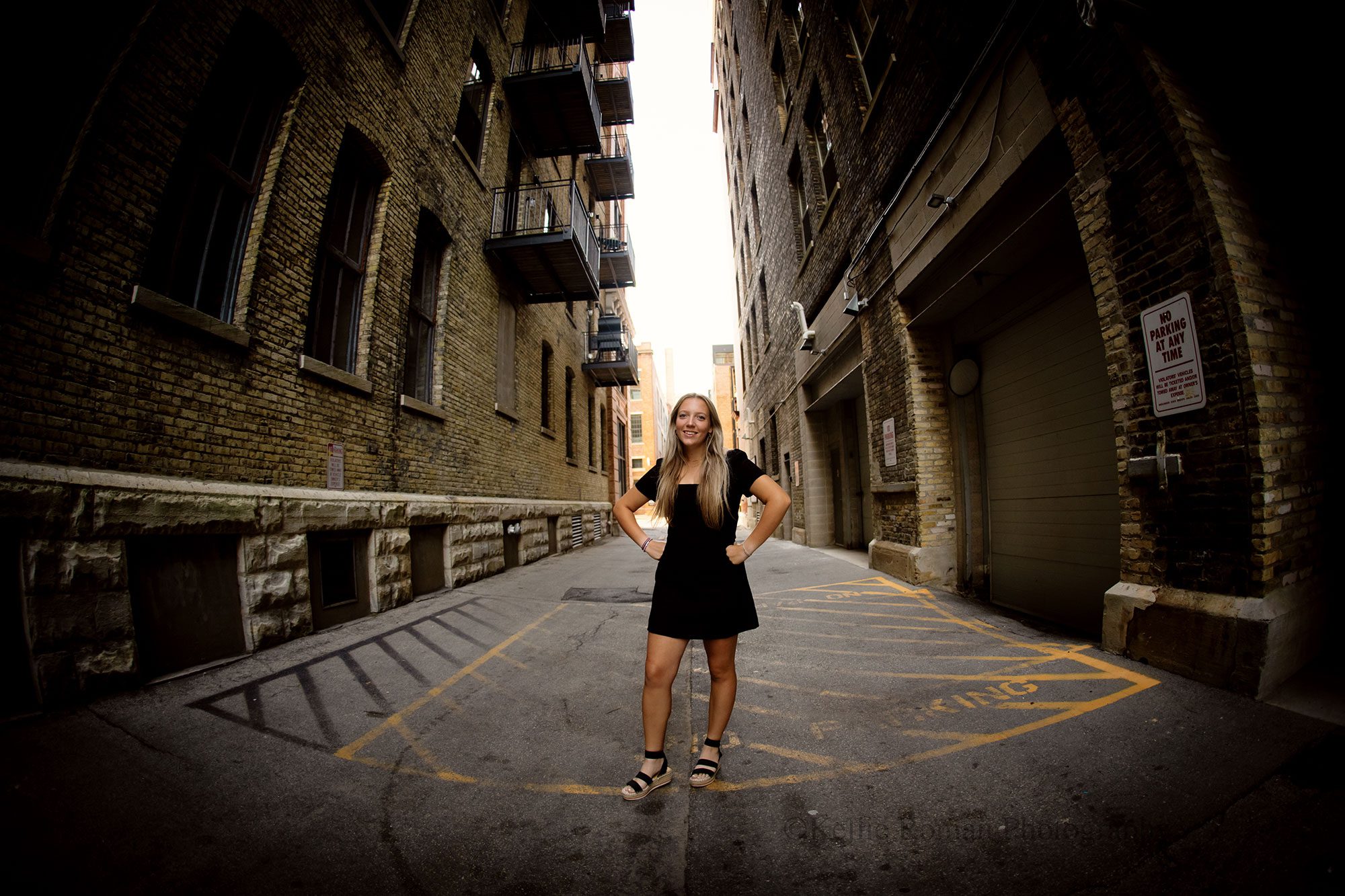 Milwaukee senior photographer. high school senior girl standing in ally in Downtown Milwaukee. she has a short black dress on and has long blond hair. she's standing with her hands on her hips.
