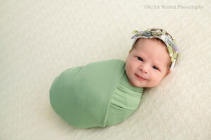 milwaukee newborn photography studio. newborn girl is awake while swaddled in a green wrap. she's laying on cream knit fabric. her eyes are open and she's smiling.