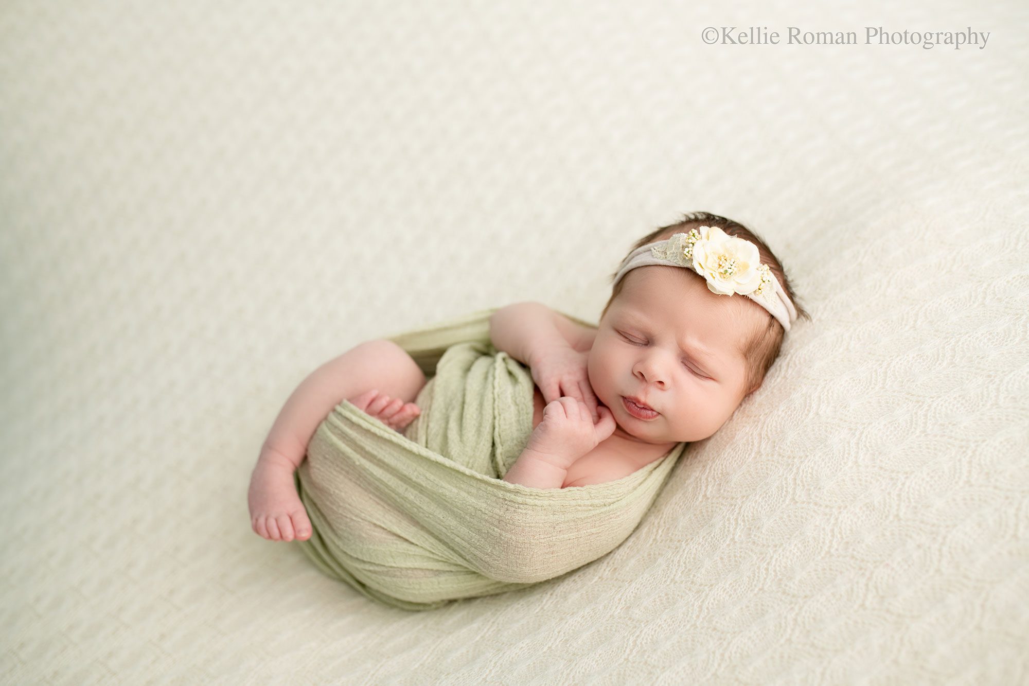 milwaukee newborn photography studio. newborn girl is sleeping onto of cream fabric. she is wrapped in a light green swaddle with her arms and legs sticking out. she has a cream floral headband on. 