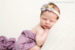 milwaukee newborn photography studio. newborn baby girl is sleeping onto of a cream knit fabric. she has a purple fabric cover her diaper. newborn is on her belly with her hand resting under her chin. baby has a purple and green floral headband on.
