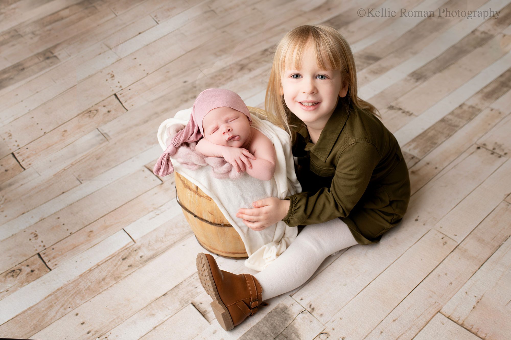 milwaukee newborn photography studio. toddler girl is sitting on floor next to wood bucket that her newborn sister is sleeping in. newborn has chin resting on arms, and a purple sleepy cap on. toddler girl has olive green dress and blonde hair. she is smiling.
