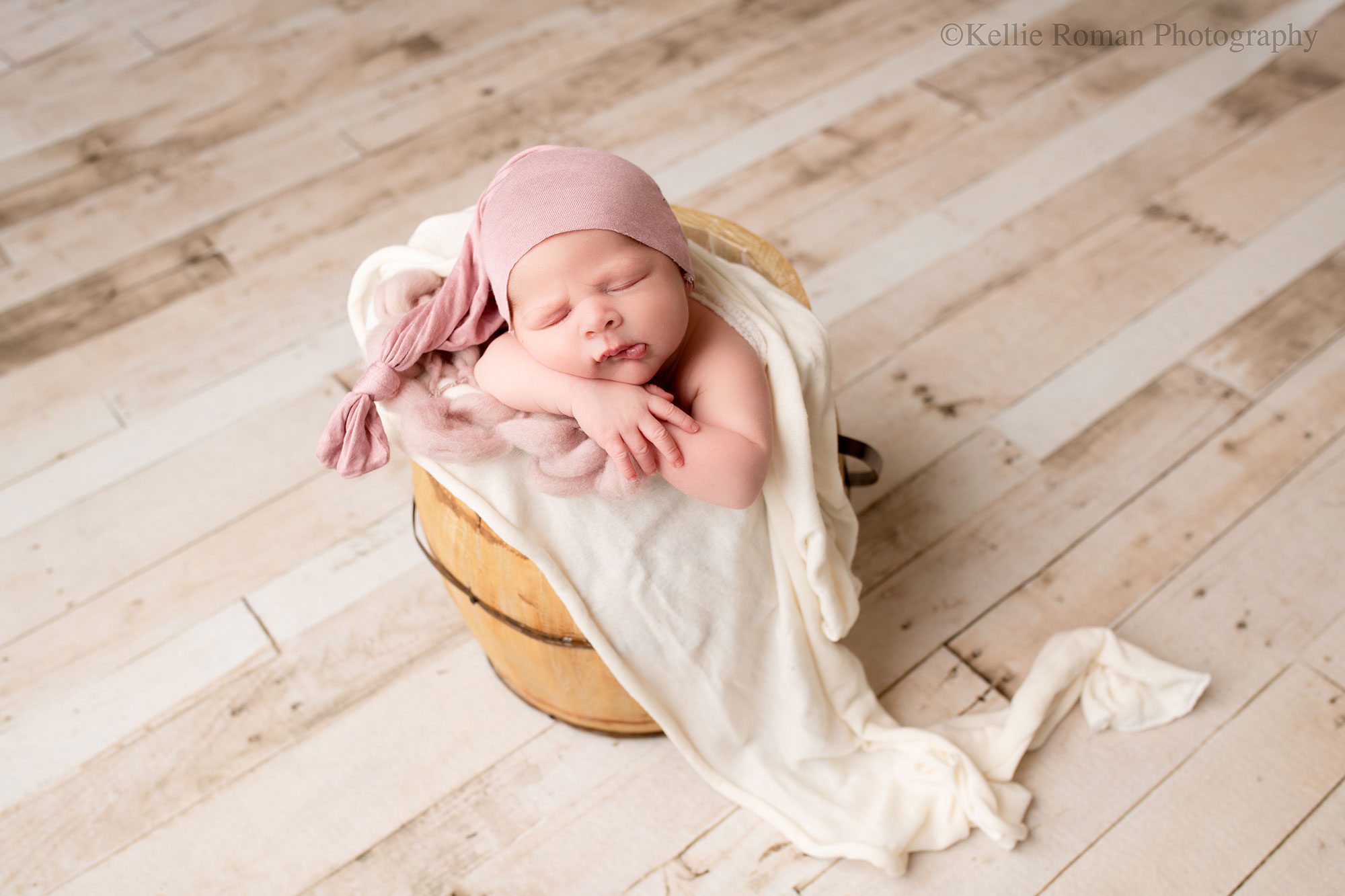 milwaukee newborn photography studio. newborn baby girl is sleeping in a wood bucket with her chin resting on her arms. the bucket is filled with light purple and cream fabric. newborn has sleepy hat on. 