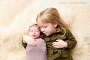 milwaukee newborn photography studio. two sisters are laying onto of cream fluffy rug. three year old girl has olive green dress on with her hand around her newborn sister while closing her eyes. newborn girl has purple swaddled around her with purple headband.