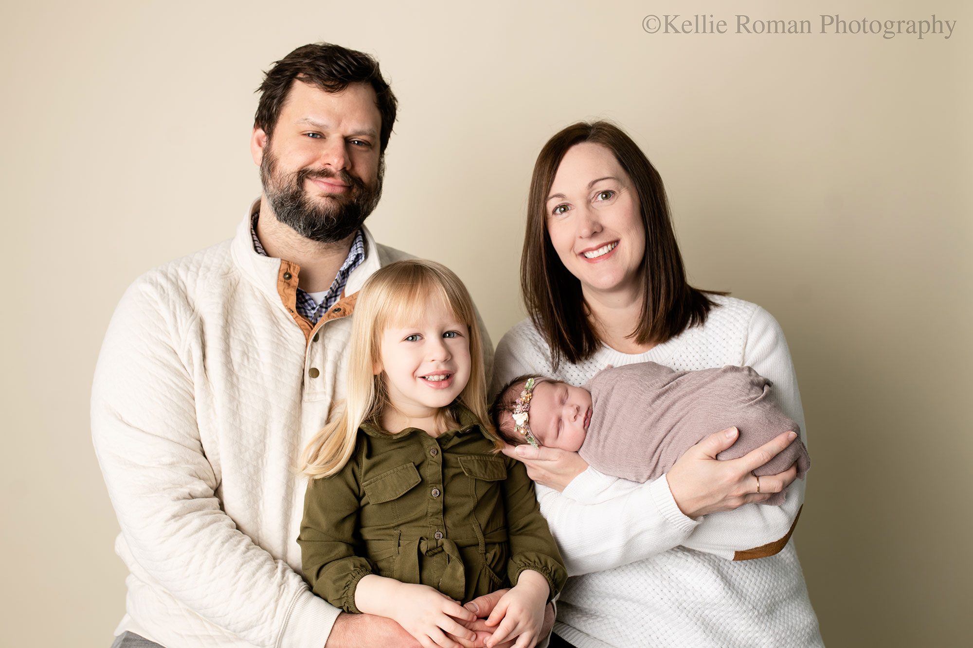 milwaukee newborn photography studio. family of four is smiling in front of a cream backdrop. dad has three year old blonde haired girl on his lap. mom is holding newborn daughter who has a floral headband on and a light purple wrap. 