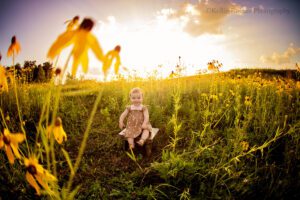 milwaukee child photographer. one year old girl sitting on wood bench. she's in a park and surrounded by yellow flowers and green grass. the sun is setting behind her so they sky is purple and blue with clouds.
