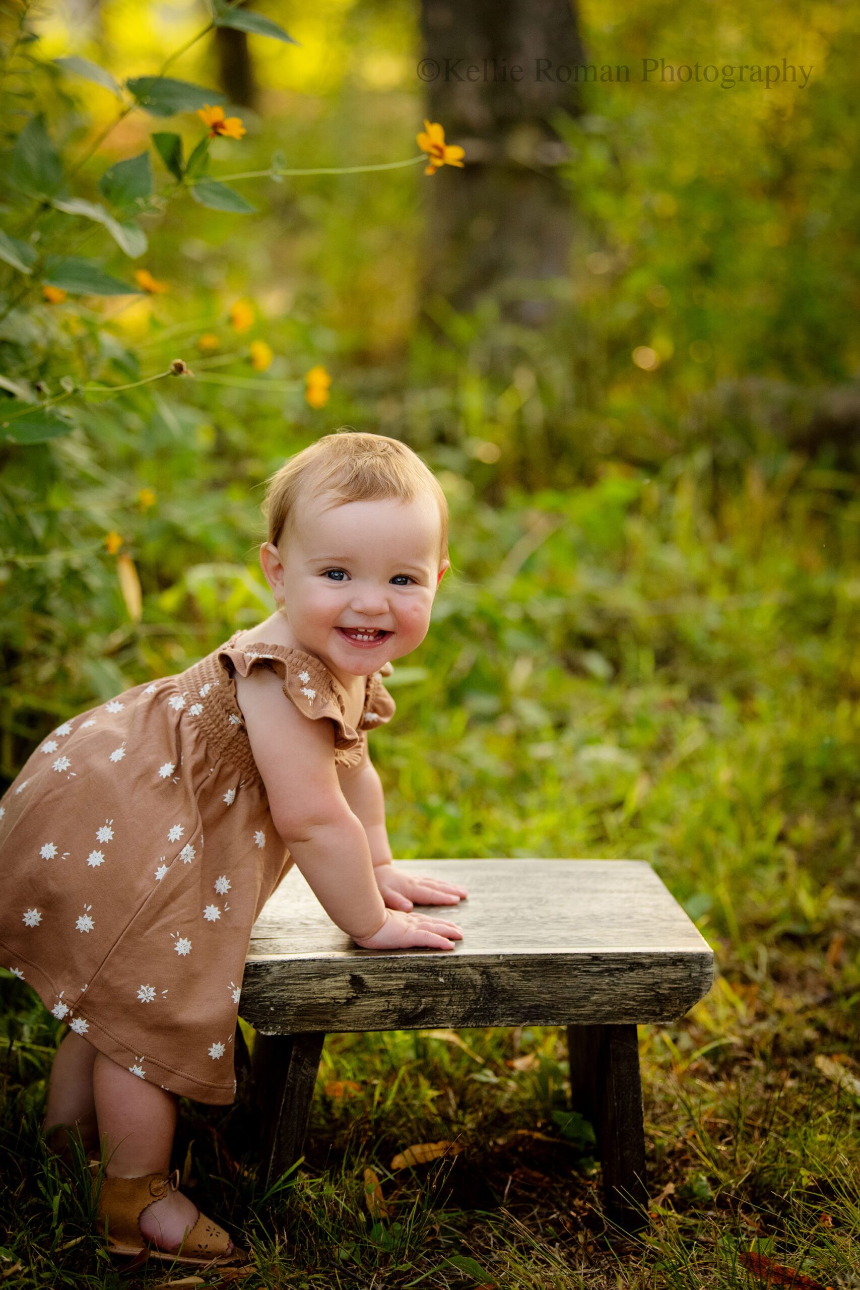milwaukee child photographer. one year old girl standing while holding onto a wood stool. girl is surrounded by green grass in a park. she has a brown dress on with white flowers and is smiling very big. 