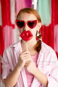 valentine mini sessions. young girl is wearing a pink and white striped shirt and red heart sunglasses. she's holding red chocolate pop up to her mouth that's in the shape of lips.