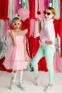 valentine mini sessions. two sisters in photo studio making goofy poses. girls are dress in pinks, teals, and whites. both girls have heart shaped glasses on. the backdrop is a valentine theme