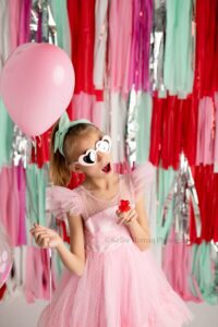 valentine mini sessions. a young girl wearing a light pink poofy tule dress is standing making a shocked face. she has pink heart sunglasses with a red heart ring pop on. she's holding onto a light pink heart balloon. the backdrop is very colorful fringe in shades of red, pink, teal, white, and silver.