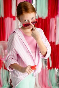 valentine mini sessions. a girl with red hair is standing in front of a fringe backdrop with colors of red, pink, teal, silver, and white. the girl has red heart sunglasses on and is pulling them down her nose with her finger. she has a white and pink striped shirt on with teal leggings.