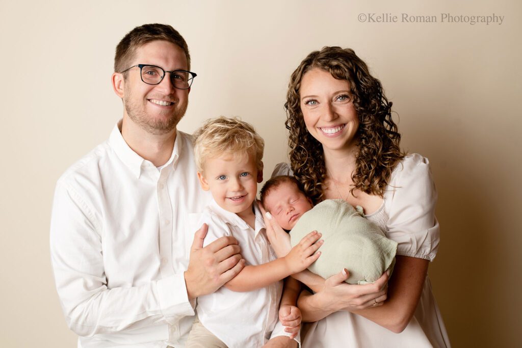 Family of four is in milwaukee photography studio. mom, dad, and two sons. one son is a three year old, and the other son is a newborn. the family is all sitting very close and smiling. the backdrop is cream and the family has shades of white one. the newborn is wrapped in a green swaddle.
