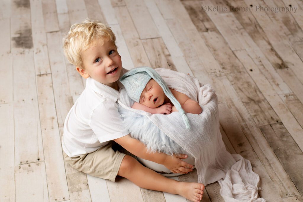 two boys in Milwaukee Photographer studio. one toddler boy is sitting on wood floor sitting next to wood bucket. newborn boy is sleeping inside of bucket with a light blue hat on. bucket is stuffed with light blue fur and white swaddle. big brother has blonde hair and a white shirt on. he's smiling at the camera.
