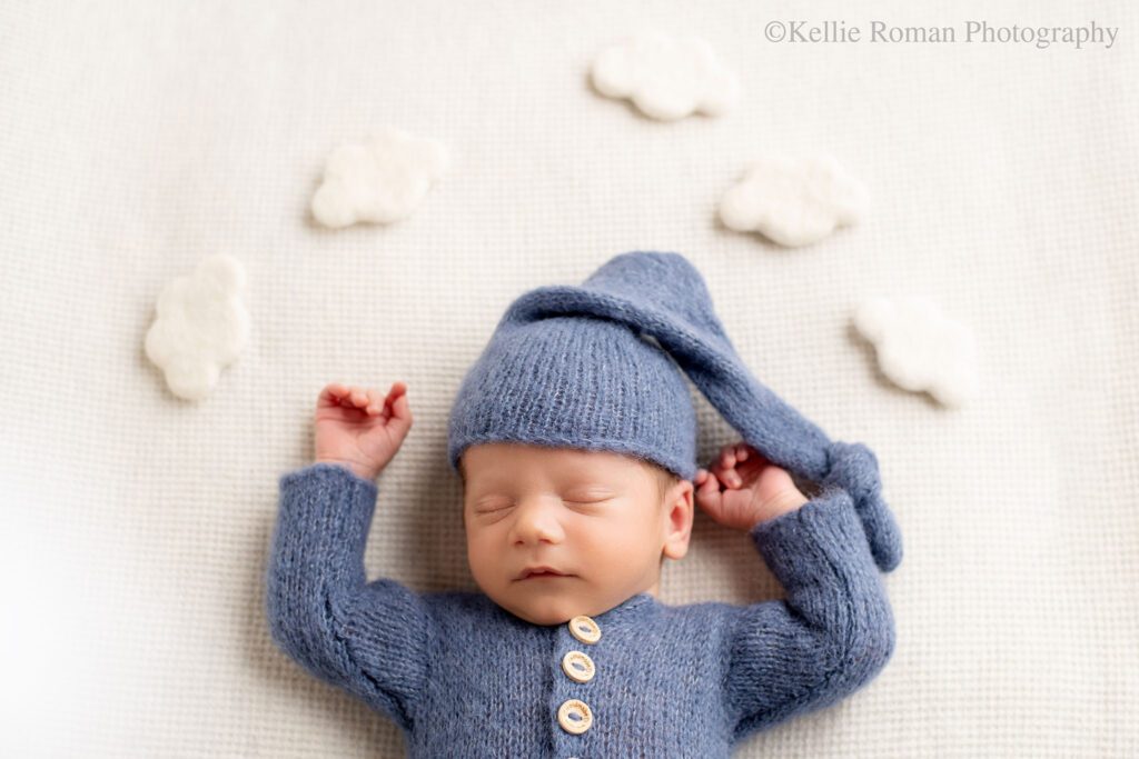 newborn is sleeping on his back onto of cream colored fabric. boy has on a blue romper with wood buttons and matching blue hat. his arms are up near his head. there are cream puffy felt clouds around his head
