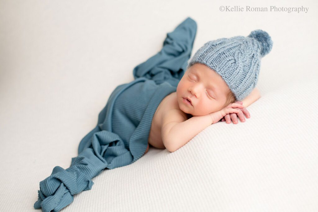 newborn baby boy is sleeping on top of cream colored fabric while a blue fabric is draped over his diaper. he has a matching blue pom pom hat on. the boy has his hands resting under his chin. 
