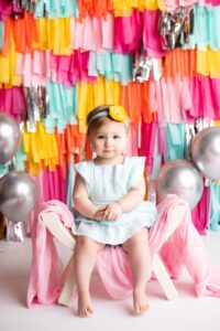 fringe cake smash milwaukee. a one year old girl sitting on a bench while smiling big at the camera. the backdrop is fun bright colors of pink, yellow, orange, teal, and silver. the one year old has a teal romper on with a yellow and teal flower headband.