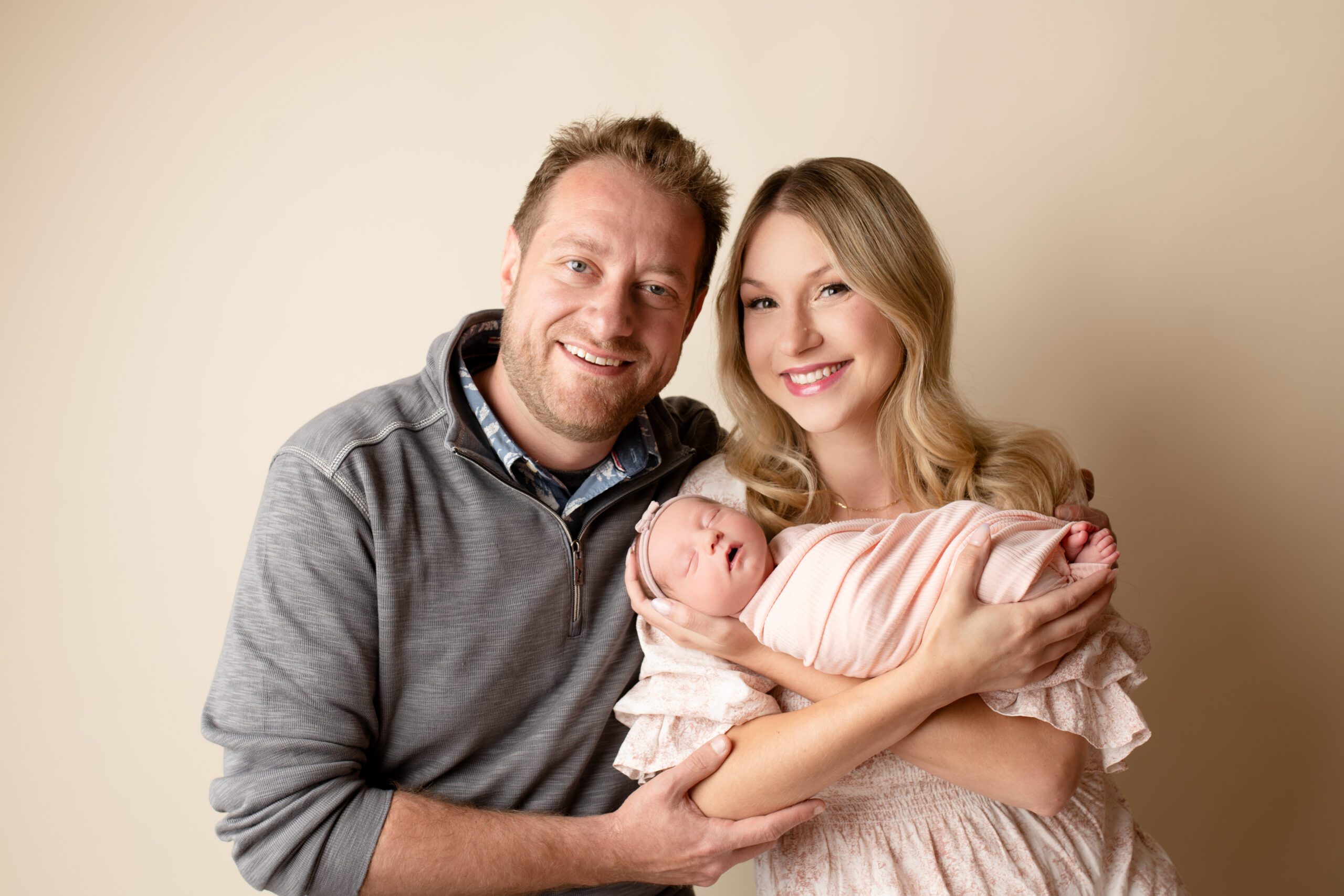 parents holding their newborn baby girl are smiling at the camera. mom has newborn in her arms and she is wrapped in a light pink fabric. 
