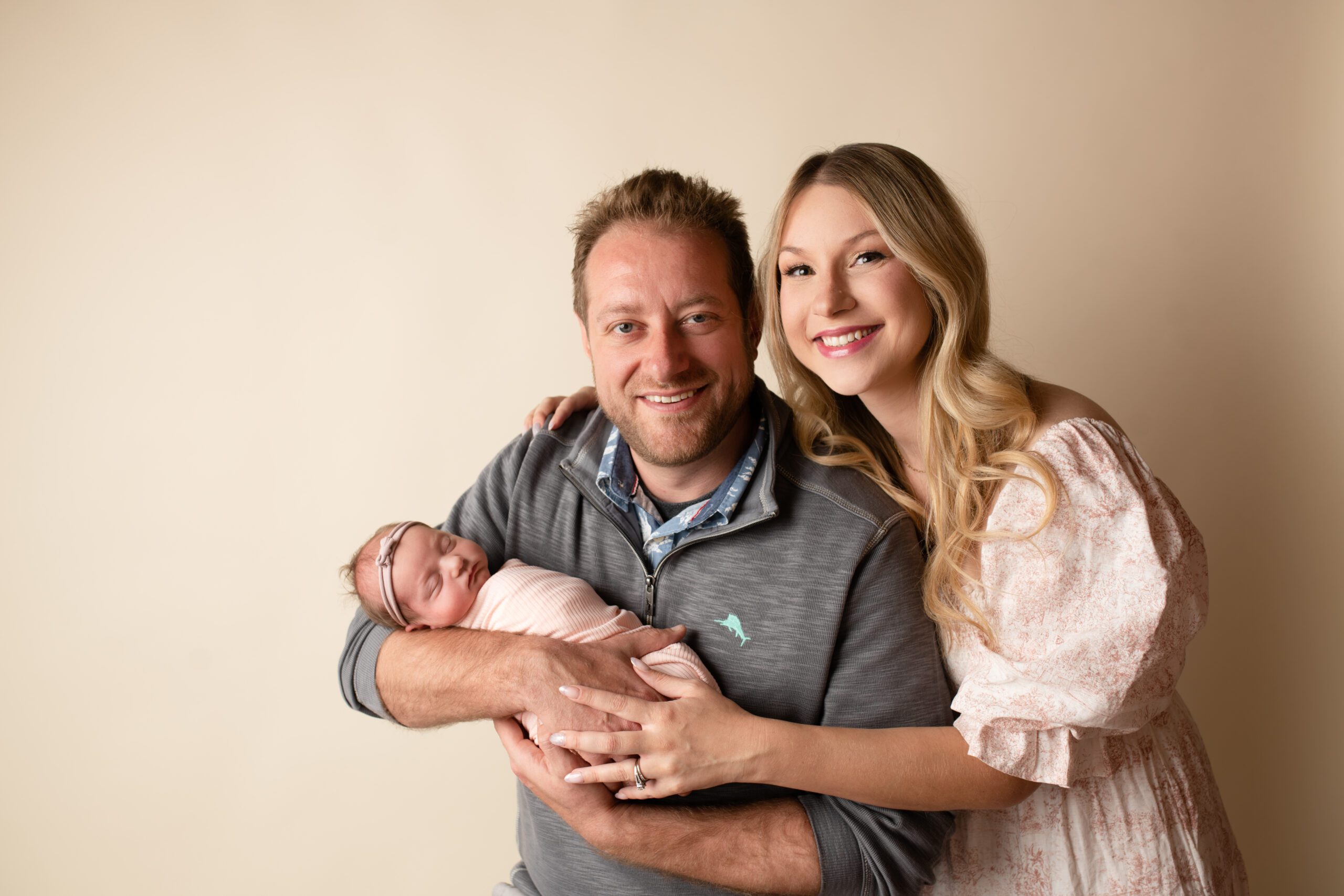 a new family of three in milwaukee photo studio. dad is holding onto newborn girl and mom is leading in towards dad. parents are smiling and baby is asleep in dads arms with light pink swaddle fabric

