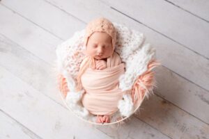 first newborn photos milwaukee. an image of a newborn baby girl in a pink swaddle. she's sleeping in a white basket on her back. the basket is filled with light pink and white fur and fabric. she has a light pink bonnet on.