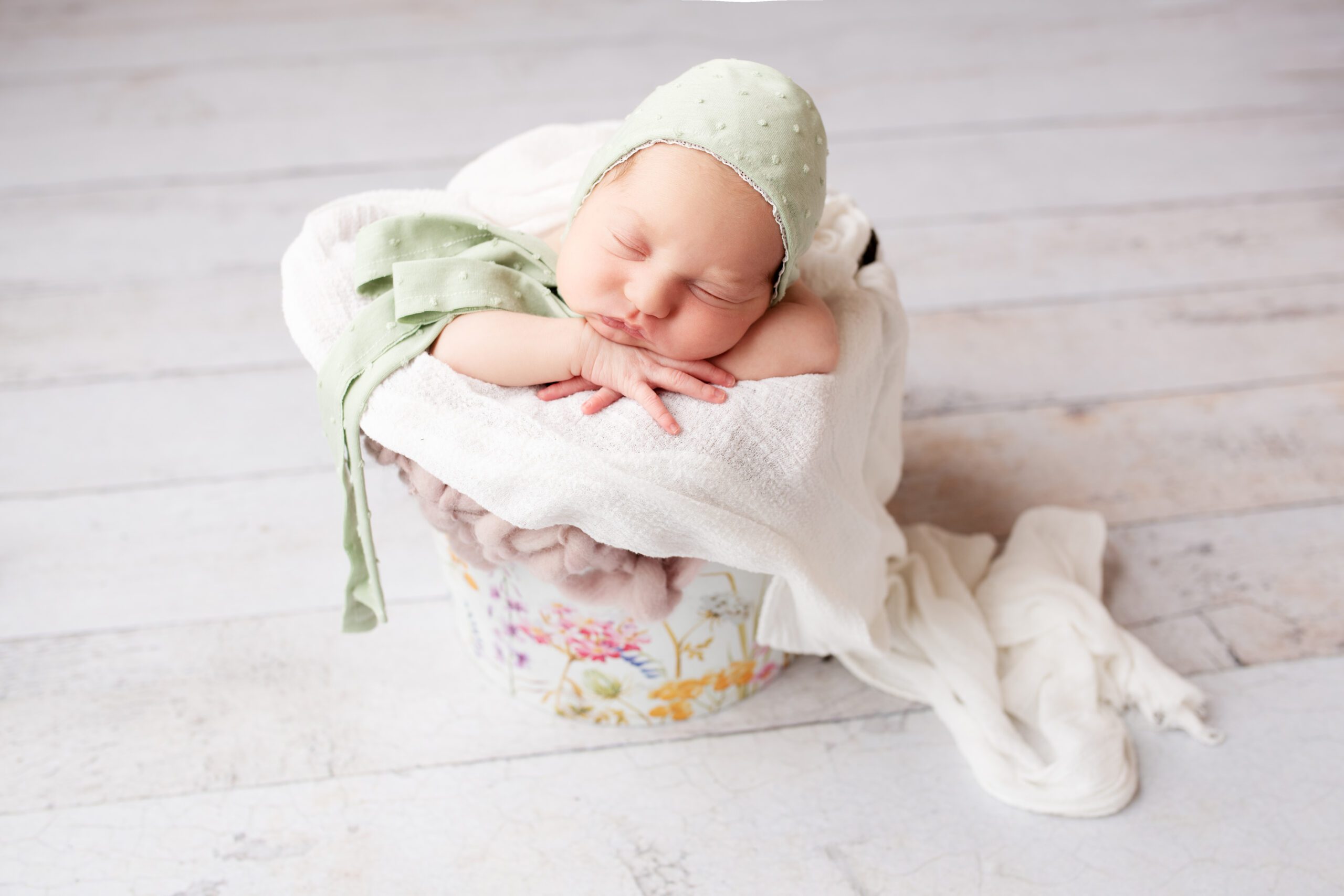 newborn girl in a bucket sleeping. her chin is resting on her hands. she is wearing a sage green bonnet. the bucket has a floral print on it and is stuffed with light purple and cream fabrics.