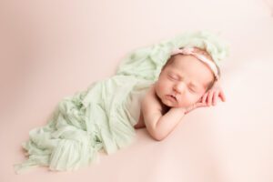first photos newborn milwaukee. a newborn girls is asleep on her tummy with her hands under her chin. she's on top of a light pink fabric with a green ruffle swaddle fabric draped over her back.