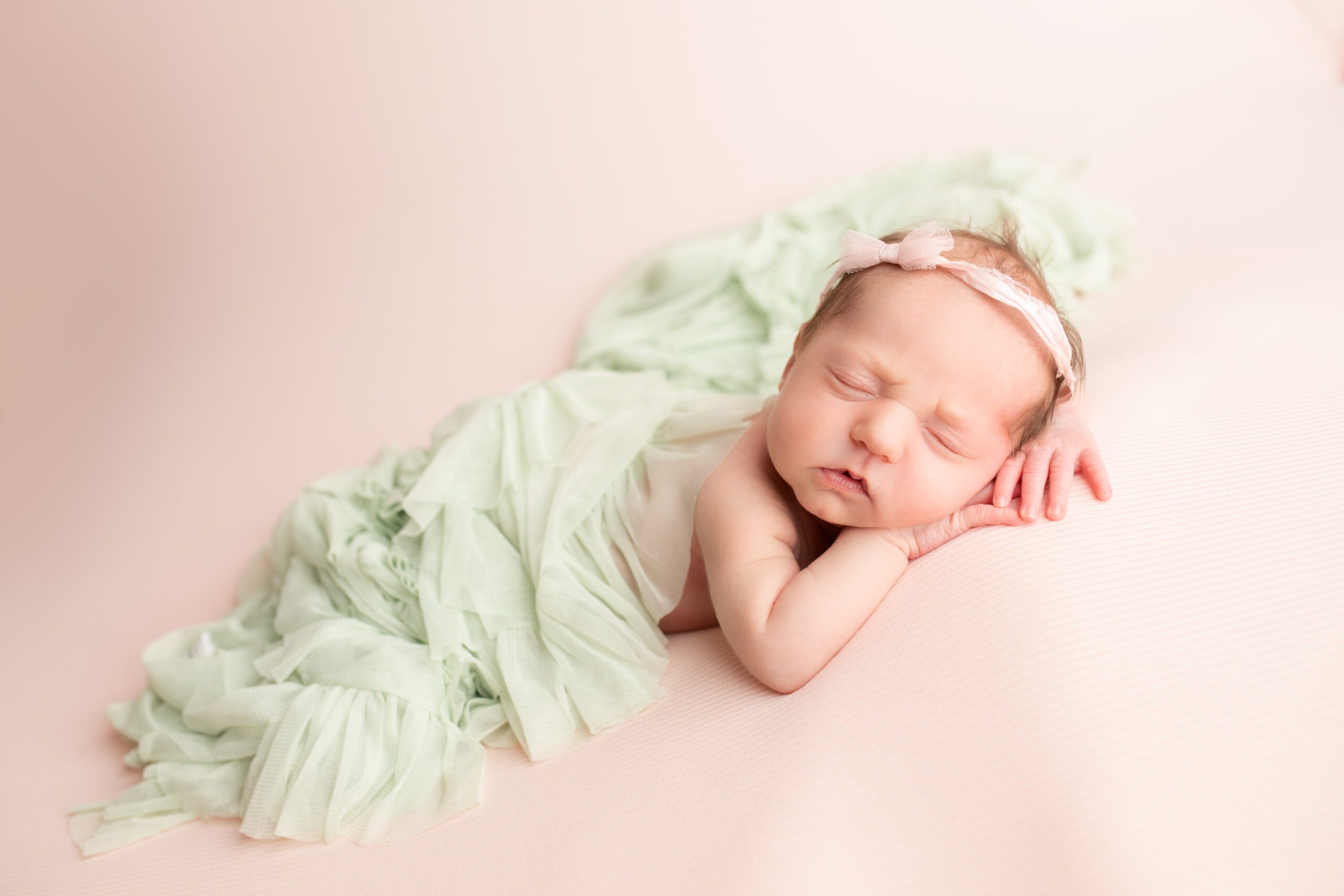newborn girl is laying on her tummy onto of a pink fabric. her diaper and back has a light green ruffle fabric draped over it. she has a light pink chiffon headband with a small bow. the newborn has her hands resting under her chin
