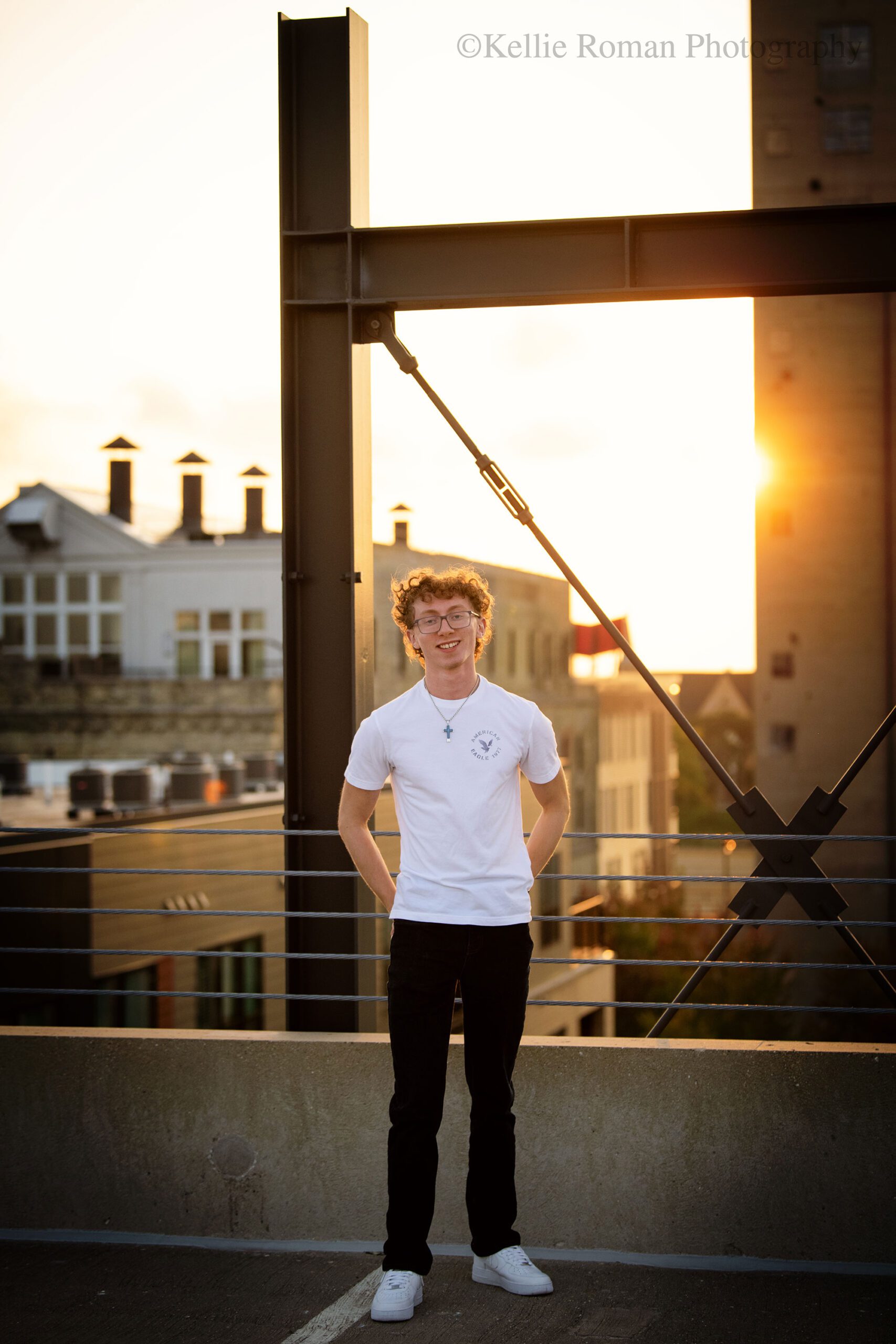 milwaukee senior photographer, milwaukee skyline. a high school senior boy is standing on rooftop of milwaukee parking garage. he has a white t shirt on with jeans and white nike shoes. he has curly hair and glasses. his hands are in his back pocket. the sun is setting behind him, and there are old buildings and metal beams in the background.
