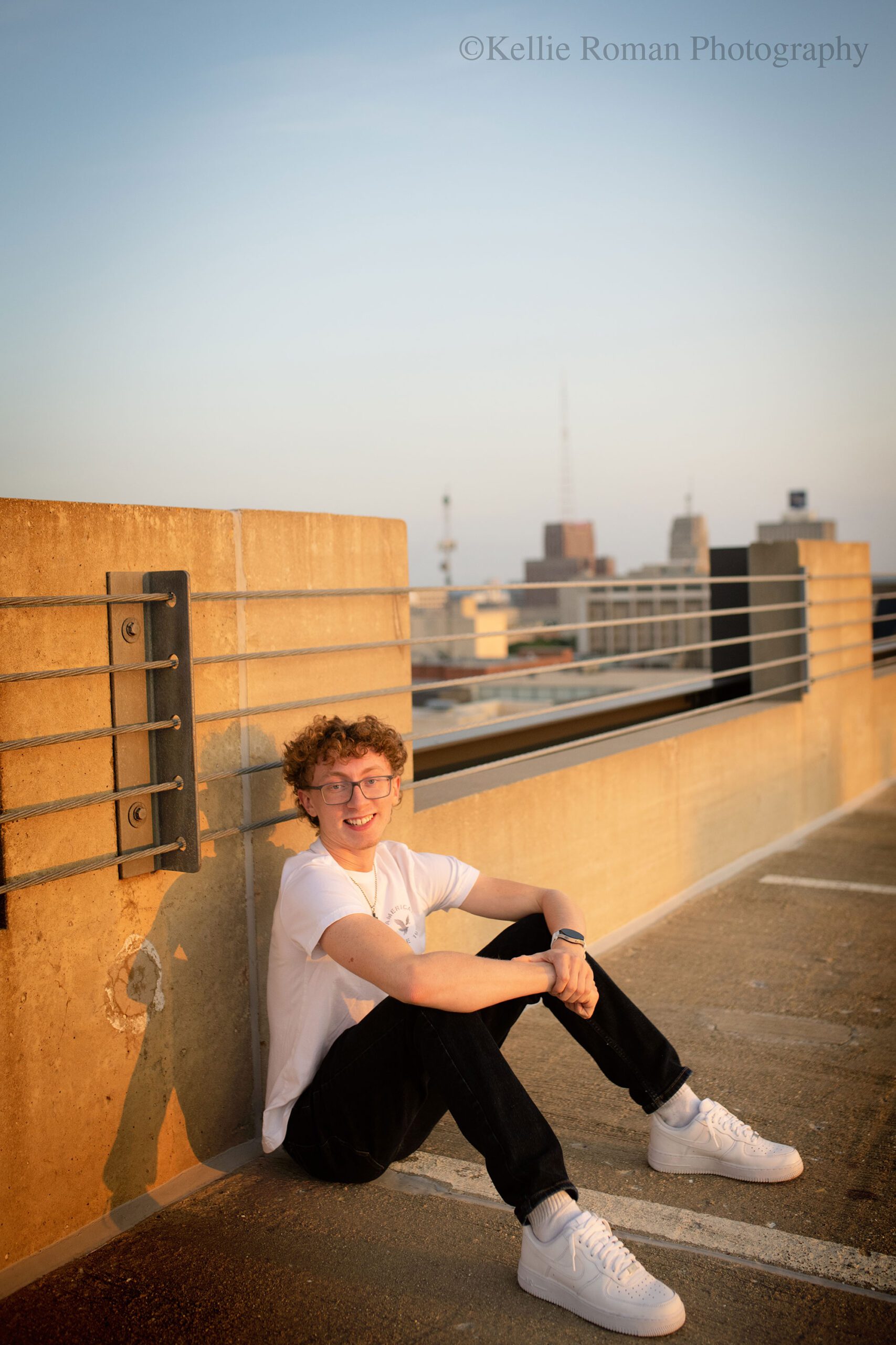 milwaukee senior photographer, milwaukee skyline. a senior boy is sitting with his back resting against concrete wall on the top of a parking garage. he has a white t shirt on with white sneakers, and jeans. he has curly hair with glasses. the sky and the tops of milwaukee buildings is in the background. 

