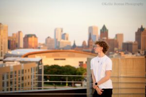 milwaukee senior photographer. high school senior boy from Greenfield is standing on rooftop of parking garage looking off in the distance. he's leaning against cable wiring, and the backdrop is the milwaukee skyline. he has a white t shirt and jeans on with glasses.