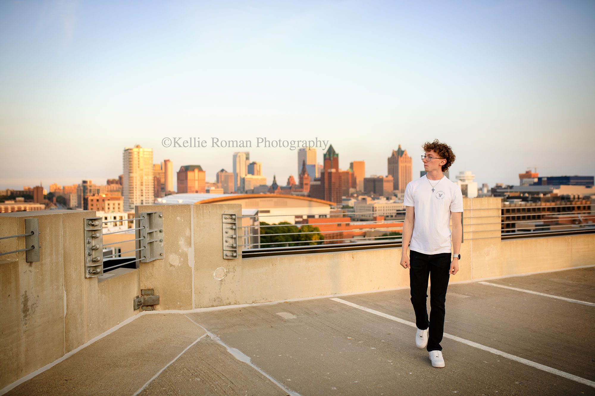 Milwaukee senior photographer, milwaukee skyline. high school senior boy is walking on rooftop of milwaukee parking garage. he's looking off to the side, wearing jeans, a white t shirt and white sneakers. the milwaukee skyline is behind him.