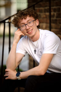 milwaukee senior photographer. high school senior boy sitting on stairs with black railing. he has elbows resting on his knees with one hand up by his face and is smiling. he has a white t shirt on with jeans, glasses and curly blonde hair.