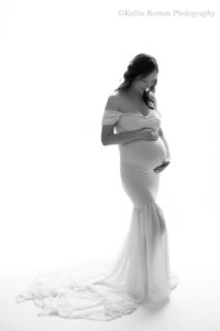 milwaukee maternity photographer. image is in black and white. women standing in front of a very bright white backdrop. she has a gown on with a long chiffon train. dress is off the shoulder. women has long black hair, and has both hands on her pregnant belly. she is looking down.