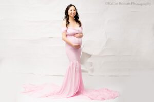 milwaukee maternity photographer. pregnant women is standing in front of a torn up white backdrop. she has a long pink dress on with a long chiffon train. women is smiling at the camera, with both hands on pregnant belly.