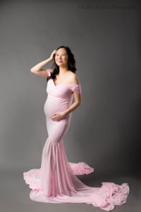 milwaukee maternity photographer. a pregnant women is standing in front of dark grey backdrop. she has a long pink gown on with train, and it's off he shoulder. she has long dark hair, and has one hand on her belly and one hand in her hair and is looking upwards.