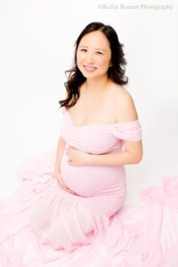milwaukee maternity photographer. a pregnant women is sitting on a white backdrop with her legs under her butt. she has a long chiffon pink dress on, that is off the shoulder. she's holding onto her pregnant belly. she is looking up and smiling at the camera.