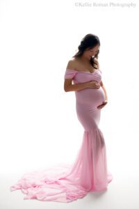 milwaukee maternity photographer. women standing in front of a very bright white backdrop. she has a light pink gown on with a long chiffon train. dress is off the shoulder. women has long black hair, and has both hands on her pregnant belly. she is looking down.