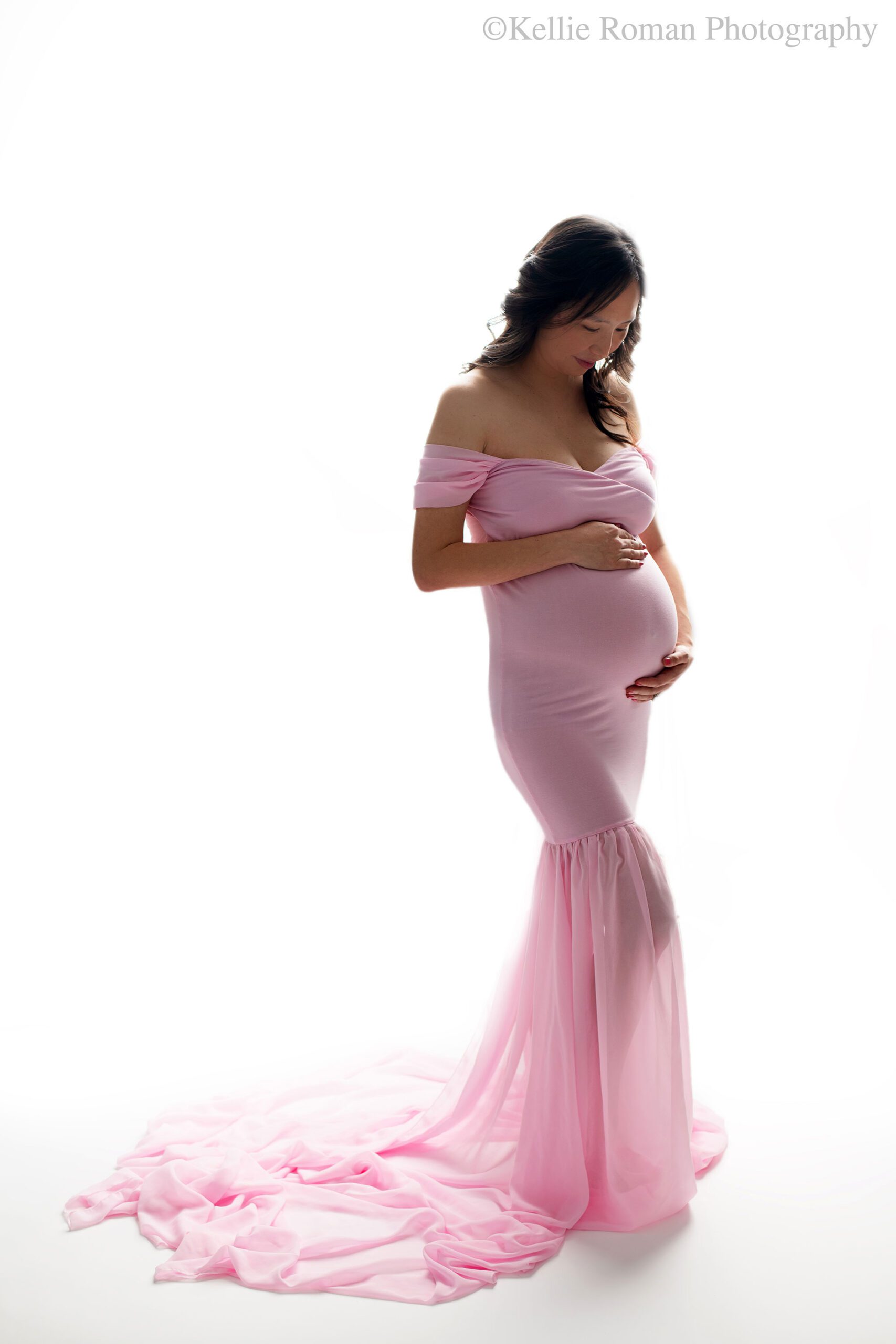 milwaukee maternity photographer. a pregnant women is standing in front of bright white backdrop holding onto her pregnant belly. she is looking down at her baby bump. she has a long pink gown on with dark black hair. 
